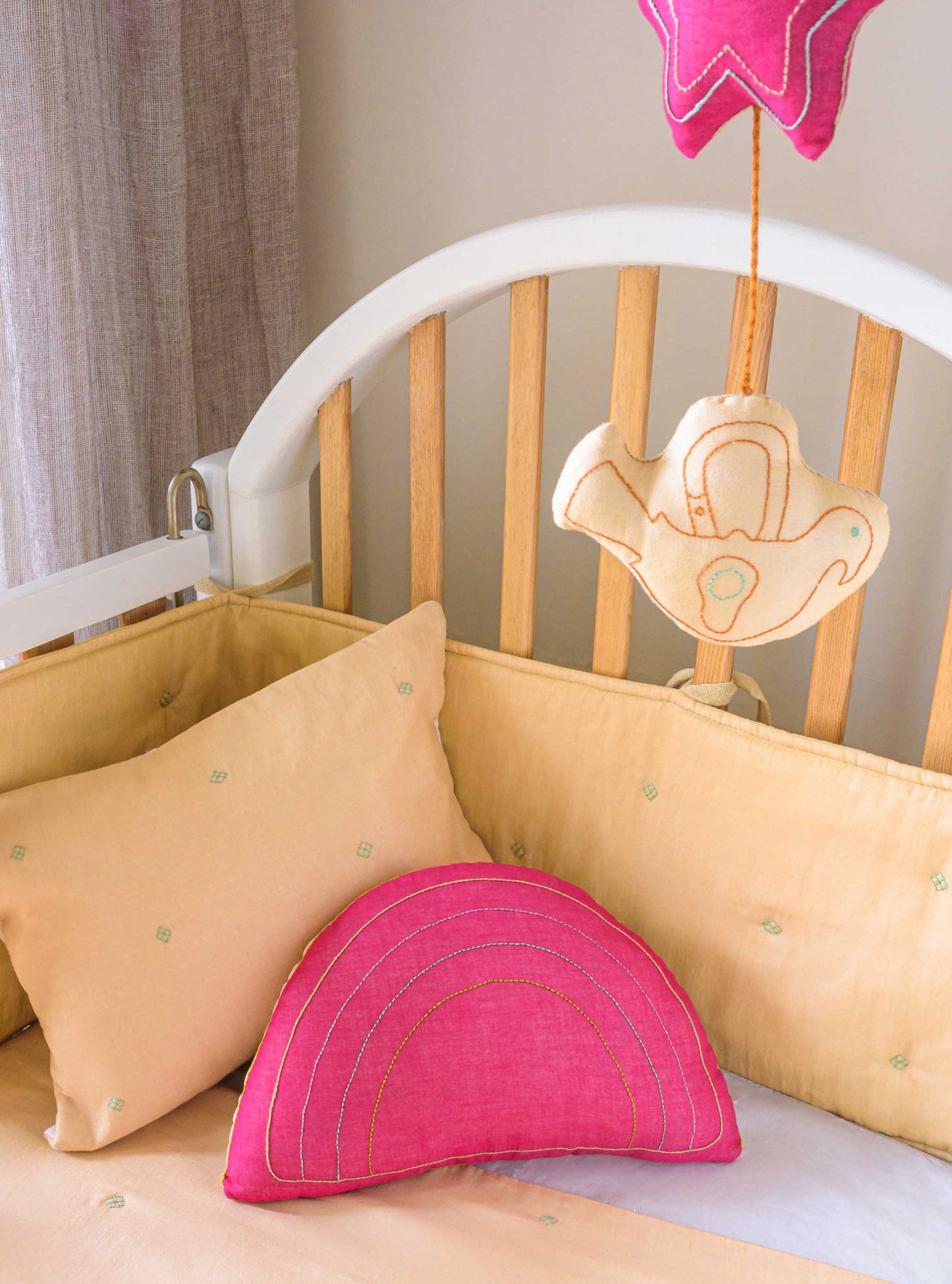 Wheat Bandook pillowcase, bumper, blanket and mobile with Fuchsia rainbow cushion all placed on a baby crib